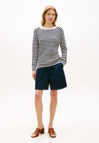 Ριγέ πουλόβερ με μακριά μανίκια σε ναυτικό μπλε και λευκό, συνδυασμένο με σορτς σε ναυτικό μπλε. Το μοντέλο φοράει καφέ mule με τακούνι, παρουσιάζοντας μια casual εμφάνιση.