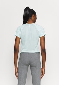 Kvinna med långt hästsvans iförd en ljusblå stickad kortärmad topp och grå tights, som står vänd bort mot en enfärgad bakgrund.