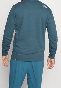 Sweatshirt teal avec un col rond, des poignets et un ourlet côtelés, et un petit logo dans le haut du dos. Texture lisse, coupe décontractée.