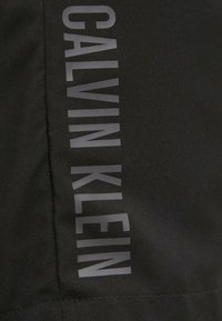 Tessuto nero con la scritta stampata "CALVIN KLEIN" in grassetto, con lettere grigie riflettenti sul lato, caratterizzato da una superficie liscia.