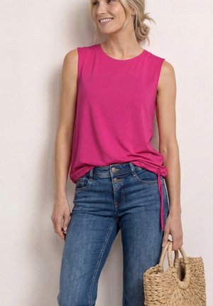 Kvinde iført en ærmeløs, knaldrød top med bindebånd i siden og blå jeans, der holder en flettet stråtaske mod en lys væg.