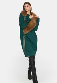 Kobieta w luźnej zielonej sukience, z brązowym futrzanym szalem na ramieniu i czarnych kozakach do kolan, pozująca z rękami blisko szala.