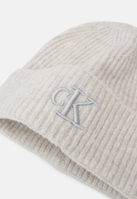 Gorro de punto gris claro con textura acanalada, que presenta un dobladillo y un logo bordado "CK" en hilo plateado en la parte frontal.