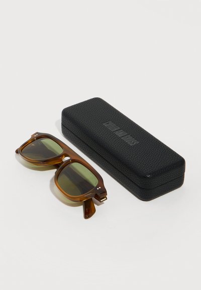 Γυαλιά ηλίου με σκελετό σε σχέδιο χελωνόκρανου και πράσινους φακούς δίπλα σε μια υφή μαύρης ορθογώνιας θήκης πάνω σε λευκή επιφάνεια.