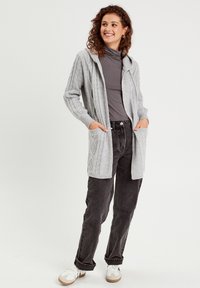 Grijze gebreide cardigan met een capuchon, voorzien van kabelbreipatronen, twee voorzakken en geribbelde manchetten. Gecombineerd met zwarte jeans en witte sneakers.