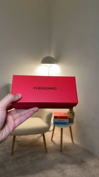Hand holding a red Ferragamo box with black text, against a background of a lit wall lamp, beige chair, and a side table with books.