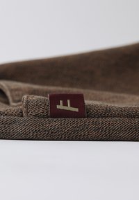 Teksturirano smeđe platno s suptilnim uzorkom, koje sadrži bordo oznaku s zelenim logotipom. Materijal ima mekan, tkan izgled.