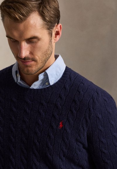 Uomo che indossa un maglione blu navy lavorato a trecce sopra una camicia azzurro chiaro con colletto, guarda verso il basso con un'espressione neutra su uno sfondo semplice.
