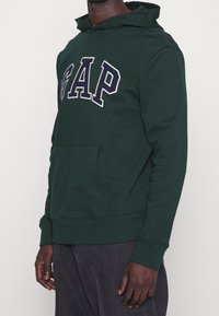 Sweat à capuche vert avec le logo "GAP" en bleu marine. Comprend une poche avant, une capuche à cordon de serrage, ainsi que des poignets et un ourlet côtelés. Texture douce.