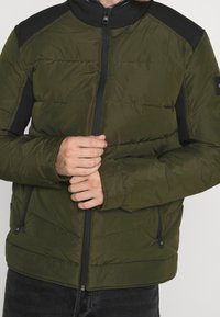 Olivengrøn polstret jakke med sorte skulderdetaljer, fuld lynlåslukning og to sidelommer med lynlås. Letvægts, quiltet tekstur.