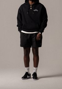 Sweat à capuche noir avec broderie "BLKVIS", associé à un short noir, des chaussettes blanches et des baskets noires avec un swoosh blanc.