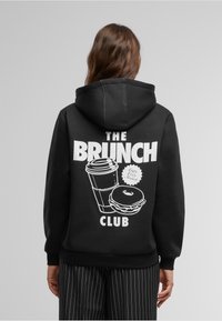 Black hoodie featuring white graphic design of a coffee cup and donut with "THE BRUNCH CLUB" text on the back. Soft fabric, casual fit.