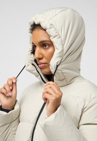 Puffy ivoorkleurige gewatteerde jas met een hoge kraag, voorzien van een zwarte rits en verstelbare trekkoorden rond de capuchon voor een veilige pasvorm.