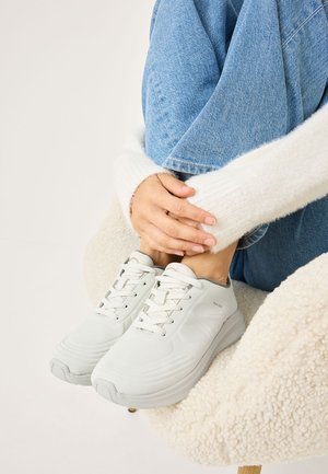 Sneaker bianche leggere con design texturizzato e lacci, abbinate a jeans in denim blu e a un maglione bianco morbido, seduto su una sedia accogliente.