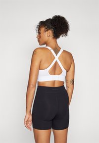 Hvit sports-bh med teksturert design og kryssede stropper, paret med svarte shorts. Har en krok-lukking på baksiden.