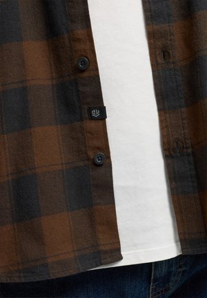 Brun og sort ternet flannel skjorte med sorte knapper, lappelomme og en hvid T-shirt nedenunder, præsenteret mod blå denim.