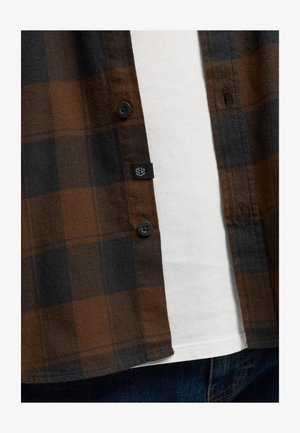 Brun og sort ternet flannel skjorte med sorte knapper, lappelomme og en hvid T-shirt nedenunder, præsenteret mod blå denim.