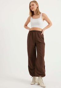 Braune weit geschnittene Hose mit elastischem Bund, ausgestattet mit einer glatten Textur und Seitentaschen; kombiniert mit einem weißen Ripp-Crop-Top und beigen Absatzstiefeln.