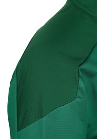 Giacca sportiva verde con tessuto testurizzato, spalle senza cuciture e sezioni lisce a contrasto. Visibili dettagli della cerniera e delle cuciture.
