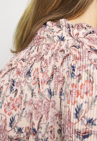 Šaty s plisovaným vzorem květin v měkké růžové látce, s řaseným výstřihem a podrobnými květinovými akcenty v modré a korálové barvě.