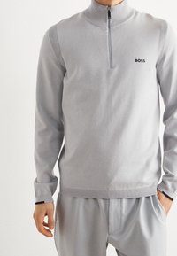 Pull à demi-zip gris clair avec des accents côtelés sur les épaules. Fabriqué en tissu doux, il présente un logo noir sur la poitrine gauche.