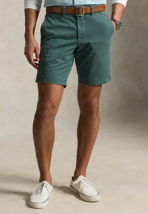 Muž v zelených šortkách, hnedom opasku, bielych teniskách a svetlomodrej košeli s vyhrnutými rukávmi, stojaci s jednou rukou v vrecku.