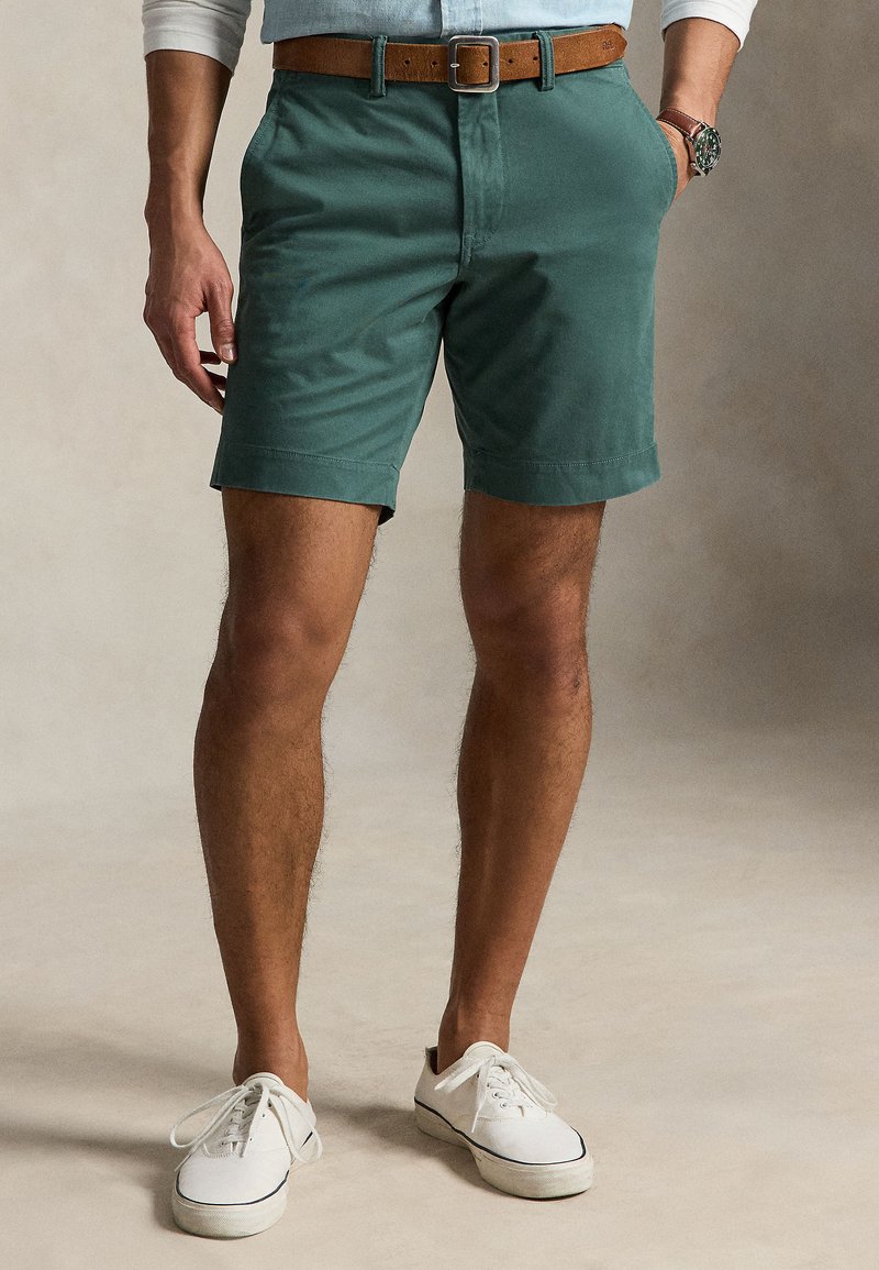 Bărbat îmbrăcat cu pantaloni scurți verzi, curea maro, pantofi sport albi și o cămașă albastru deschis cu mânecile suflecate, stând cu o mână în buzunar.