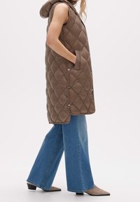 Brun vadderad ärmlös väst med huva, som har sidofickor och tryckknappar, kombinerad med vida blå denimbyxor.