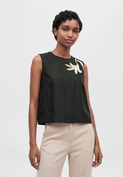 Un mannequin porte un haut noir sans manches avec un détail floral couleur crème et un pantalon beige clair, et se tient devant un fond uni.