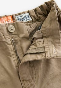 Pantalones cortos caqui con cierre de cremallera, cintura elástica, detalle de botón y bolsillos cosidos. Presenta logotipos de Superdry en la cintura.