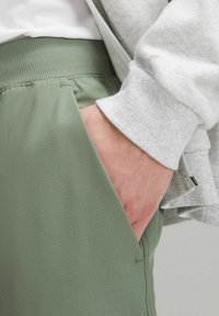 Světlé zelené sportovní kalhoty s žebrovaným pasem, vybavené bočními kapsami a hladkým, matným povrchem, v kombinaci se šedou mikinou.