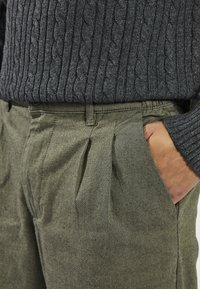 Pull gris en maille torsadée associé à un pantalon plissé vert olive avec poches latérales et tissu texturé.