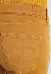 Yellow cotton pants featuring a back pocket with zigzag stitching and a metal logo accent. Smooth texture with a tailored fit.