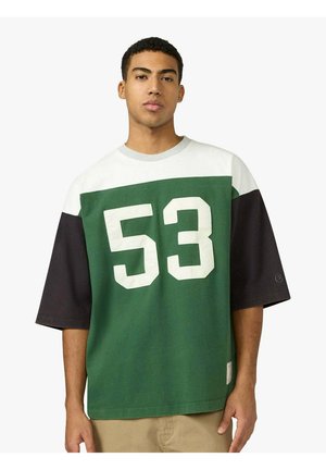 Giovane uomo che indossa una maglia oversize verde, bianca e nera con il numero 53 stampato in grande bianco sulla parte anteriore, in piedi contro uno sfondo semplice.