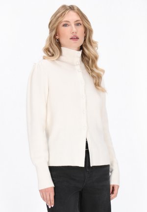 Femme aux cheveux blonds ondulés portant un cardigan blanc boutonné avec un col montant côtelé et un pantalon noir sur un fond blanc.