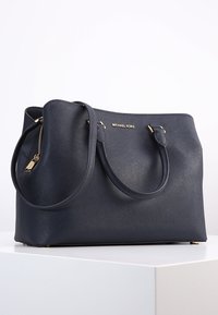 Borsa a tracolla in pelle blu navy con due manici, dettagli in metallo dorato e finitura testurizzata. Presenta una chiusura con zip e logo brandizzato.