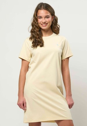 Fiatal nő hosszú, hullámos hajjal, mosolyogva, halvány bézs színű, rövid ujjú pólóruhában, semleges háttér előtt.