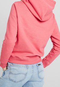 Sudadera con capucha rosa, con un bolsillo tipo canguro, puños y dobladillo acanalados. Llevada con jeans de mezclilla azul claro que presentan costuras visibles.