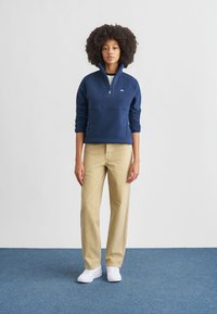 Marineblå fleecegenser med glidelåskrage, kombinert med lyse beige bukser og hvite joggesko. Enkel design og komfortabel passform.