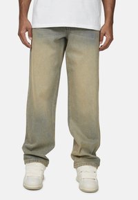 Jeans en denim délavé, coupe relaxée, jambes fuselées, couleur fanée avec des accents plus foncés. Portés avec des baskets blanches et un t-shirt blanc simple.