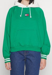 Sudadera verde con capucha blanca, que presenta un parche con logo. Puños acanalados con rayas, corte holgado y un bolsillo canguro en la parte frontal.