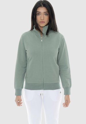 Femme aux longs cheveux foncés portant une veste verte sauge zippée et un pantalon blanc, debout devant un fond gris clair.