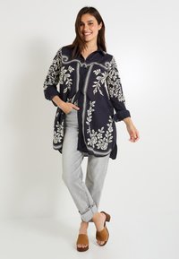 Vestido camisero azul marino con bordado floral blanco y detalles de patrón, con cierre de botones, mangas largas y un corte relajado. Combinado con jeans grises.