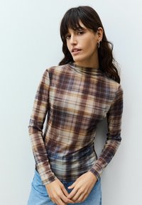 Dlouhý rukáv, fitted top z průhledného materiálu s hnědým a béžovým kostkovaným vzorem a vysokým výstřihem. Kombinováno se světle modrými džíny.
