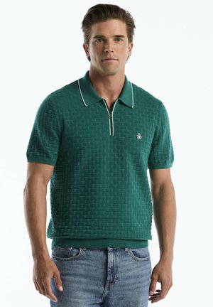 Mann trägt grünes, strukturiertes kurzärmliges Poloshirt mit weißem Besatz und blaue Jeans, steht vor einem schlichten hellen Hintergrund.