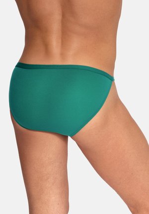 Man die effen groene zwembroek draagt, gezien van achteren, met zicht op de onderrug, heupen en bovenbeen tegen een effen achtergrond.