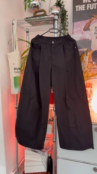 Crne široke hlače s elastičnim pojasom, u kojima su dva bočna džepa i zatvarač na gumb. Glatki materijal sa sjajnim završetkom.