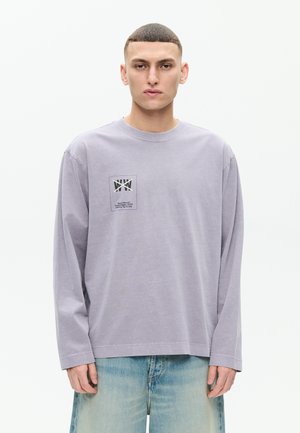 Mladý muž s krátkymi vlasmi, oblečený v svetlofialovej košeli s dlhými rukávmi s malým čierno-bielym grafickým motívom na hrudi a vybledlými modrými džínsami.