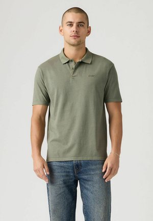 Polo vert olive avec une texture douce, patte de boutonnage à trois boutons et petit logo brodé. Porté avec un jean bleu en denim.