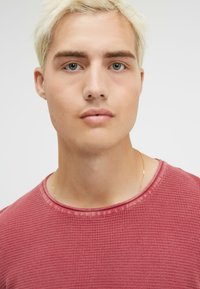 Rotes T-Shirt mit Waffelstruktur und Rundhalsausschnitt. Sichtbare Nähte und ein subtiler Glanz. Mann trägt eine zarte goldene Halskette.